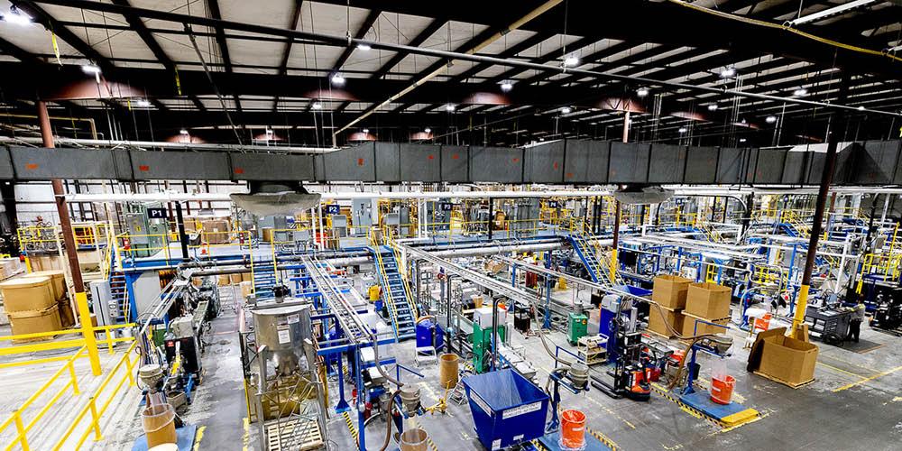 Large industrial manufacturing facility with machinery, conveyor belts, storage containers, and overhead lighting, viewed from an elevated perspective.