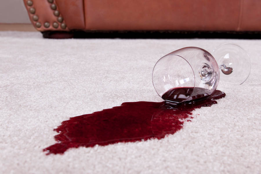 A glass of red wine tipped over on a light-colored carpet, spilling wine and creating a large stain.