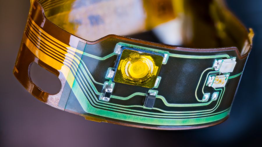 Close-up of a flexible electronic circuit with a yellow LED and various components on a transparent, curved surface.