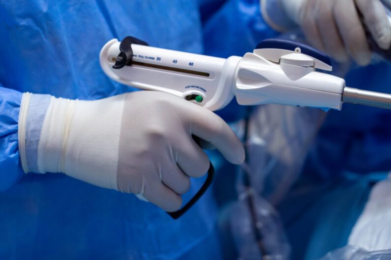 A gloved hand holds a surgical stapler during a medical procedure, with a blue sterile drape and another gloved hand visible in the background.