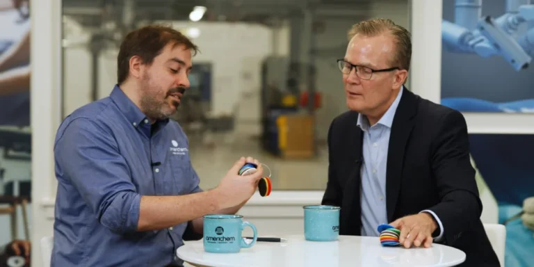 Two men sit at a table with blue mugs, one showing colored cable samples to the other in a professional setting.