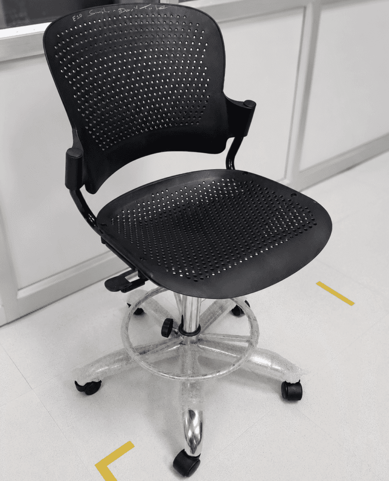 A black perforated office chair with armrests, adjustable height, and five caster wheels is positioned on a tiled floor near a wall with a glass panel.