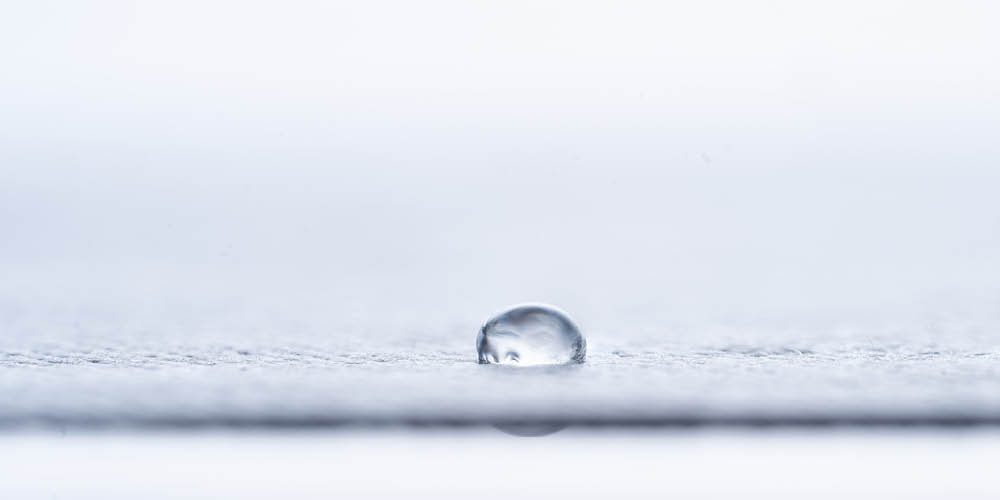 A single water droplet rests on a smooth, light-colored surface with a blurred background.