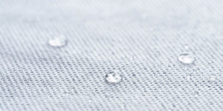 Close-up of water droplets resting on a light grey, textured fabric surface, indicating that the material is likely water-resistant or waterproof.
