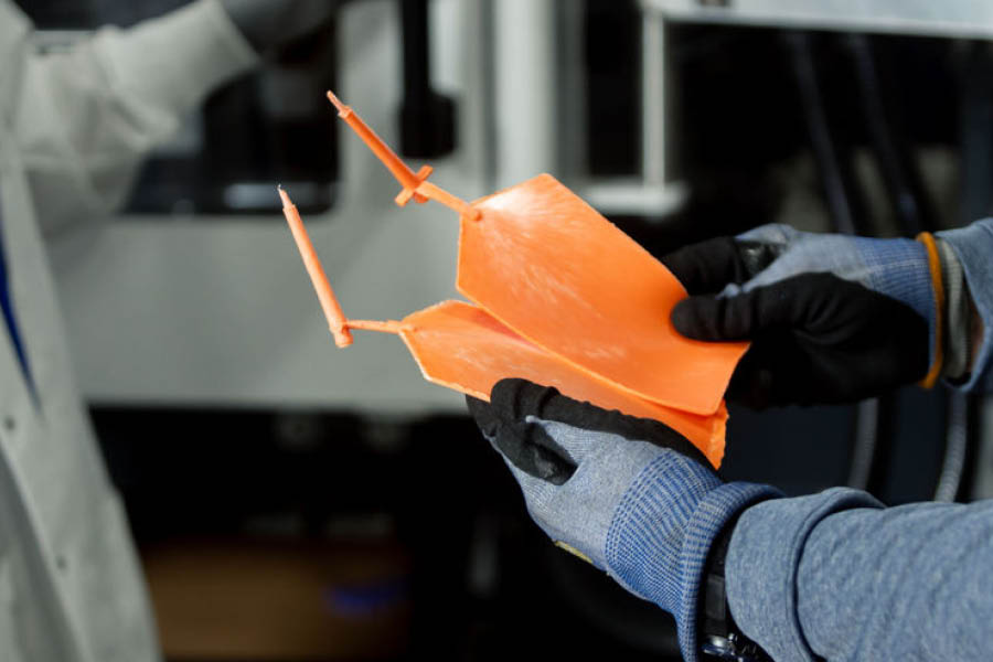 Gloved hands holding an orange 3D-printed object with angular wings and upright fins; a person in a lab coat stands in the background.