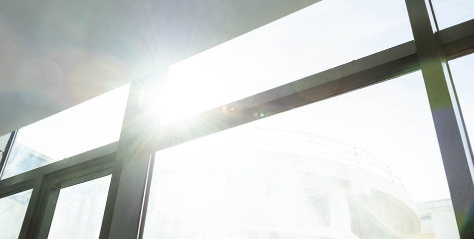 Bright sunlight shines through large glass windows, creating lens flare and illuminating part of a modern building structure outside.