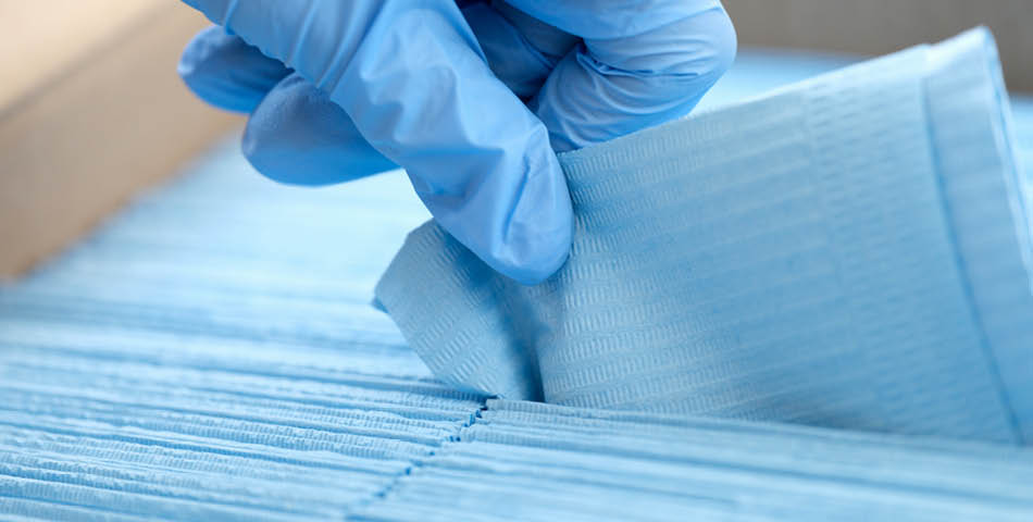 A gloved hand picks up a blue disposable face mask from a stack of similar masks.
