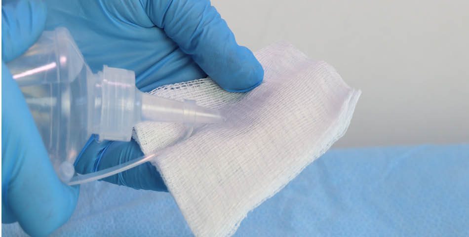 A gloved hand sprays liquid onto a piece of gauze, which is held over a blue medical drape.