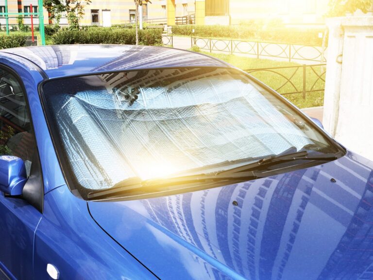 Blue car parked outside with a reflective sunshade placed inside the windshield to block sunlight, with bright sunlight shining on the front of the vehicle.