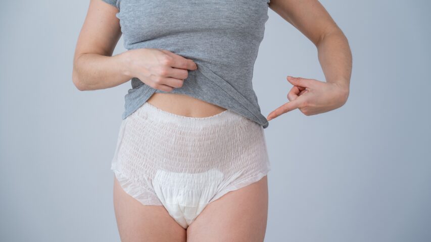 Person wearing a gray shirt and adult diaper, lifting shirt slightly and pointing at the diaper, against a plain background.