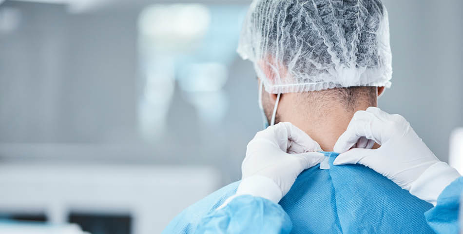 A medical professional in scrubs and a hair net is having their gown secured by another person wearing white gloves in a clinical setting.