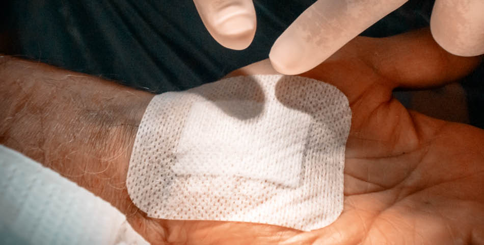 A gloved hand adjusts a white medical bandage on a person’s wrist.