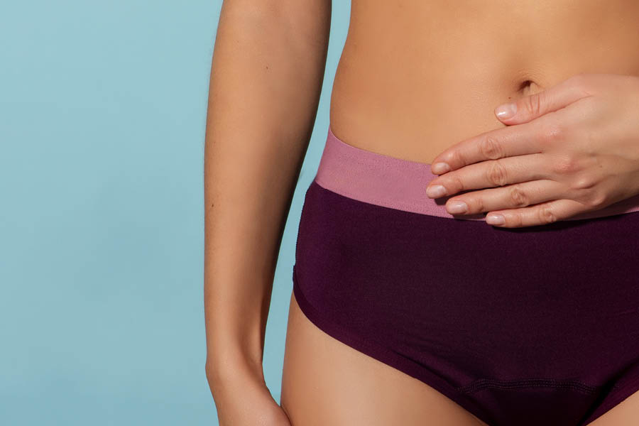 A person wearing purple underwear stands against a blue background with one hand resting on their lower abdomen.