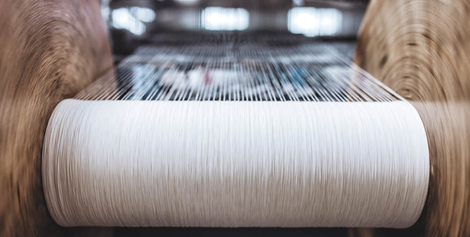 Close-up of a textile machine with white threads being woven between two large spools in a factory setting.