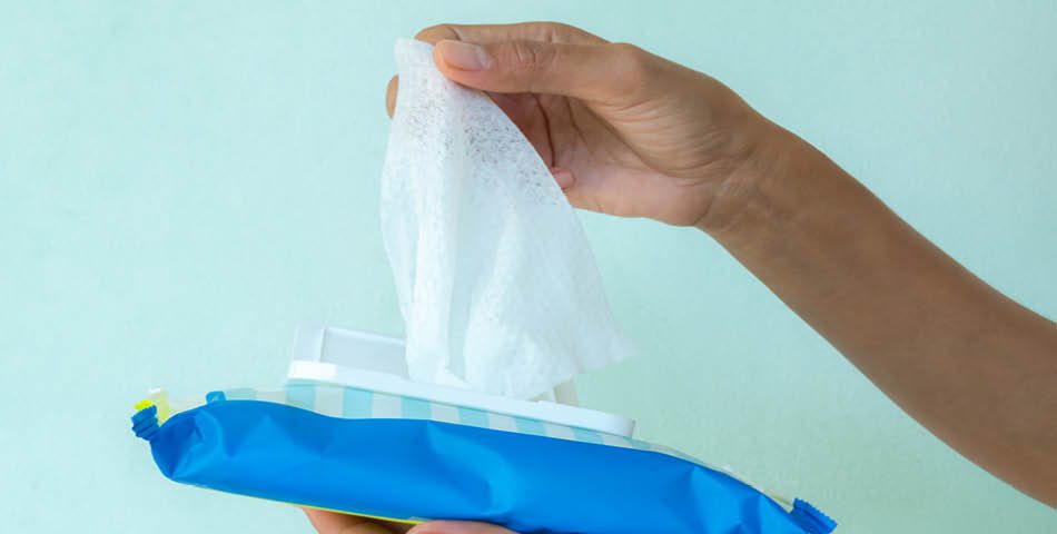 A hand pulls a white wet wipe from an open blue plastic package against a light green background.