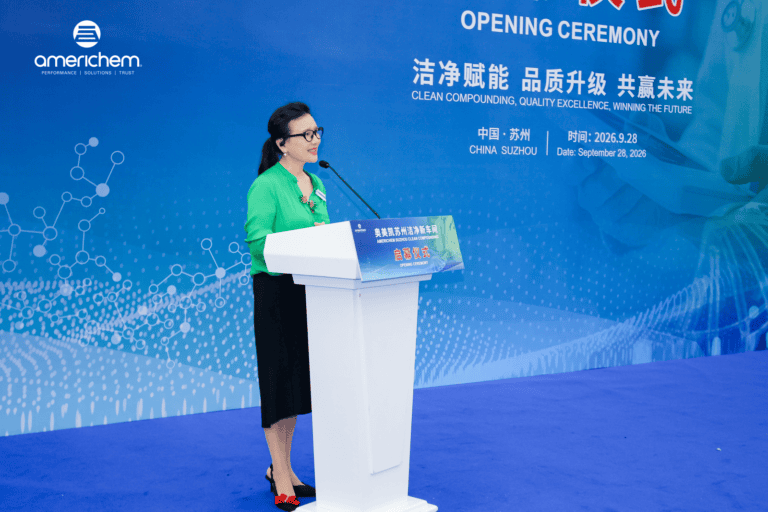 A woman in a green blouse stands at a podium giving a presentation in front of an Americhem opening ceremony backdrop.